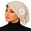 A woman shown in profile wearing a beige hijab with rhinestone embellishments and a cream fabric flower accent, paired with a black turtleneck against a plain white background.