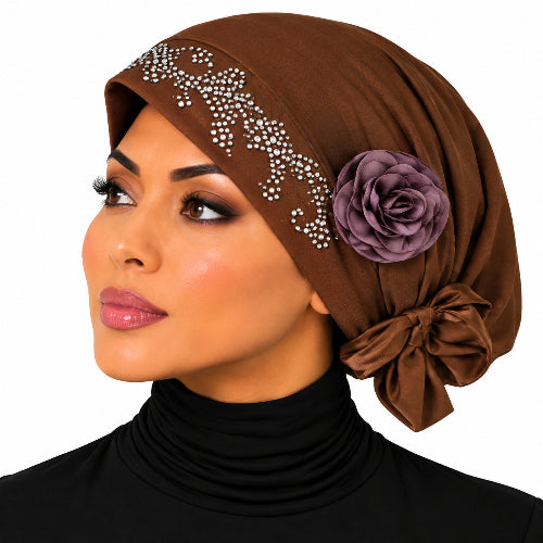 A woman wearing a brown hijab with silver rhinestone embellishments, a purple fabric rose, and a side bow, paired with a black turtleneck against a white background.