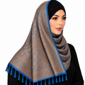A woman wearing a patterned hijab with blue tassels, styled with a black headscarf underneath, against a solid white background.