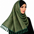 A woman wearing a green patterned hijab with tassel trim, styled over a black headscarf, against a plain white background.