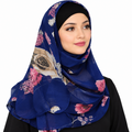 A woman wearing a royal blue chiffon hijab with a large floral print featuring pink, beige, and white roses, styled with a black undercap against a plain white background.