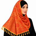 A woman wearing a bright orange and yellow patterned hijab with tassels, styled over a black headscarf and black top, against a solid white background.