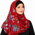 A woman wearing a red chiffon hijab with a blue, pink, and white floral print, styled over a black headscarf and a black top against a plain white background.