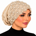 Woman modeling a beige embellished turban with floral lace, pearl beads, and sparkling details on a white background.
