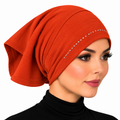 A woman wearing a bright rust-orange hijab with a curved line of small silver rhinestones across the front, shown in a close-up profile against a plain white background.