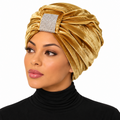 A woman wearing a gold velvet turban headwrap with a rhinestone-embellished square accent on the front, paired with a black turtleneck against a plain white background.