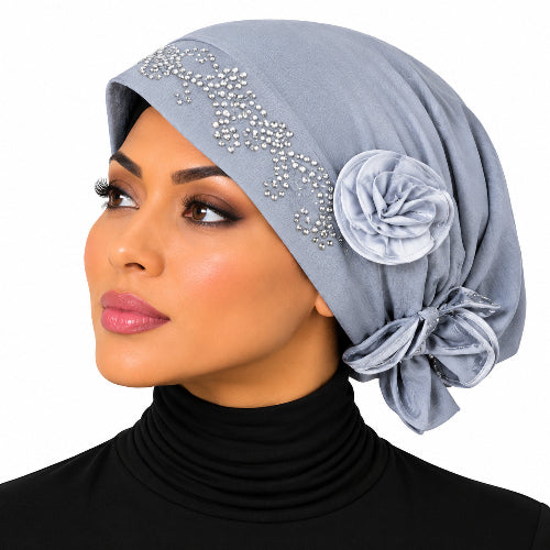 A woman shown in profile wearing a light blue-gray hijab with silver rhinestone embellishments and a large fabric flower accent on the side, against a plain white background.