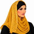 A woman wearing a mustard yellow hijab with rhinestone embellishments on the side panel, paired with a black undercap and black top, against a plain white background.