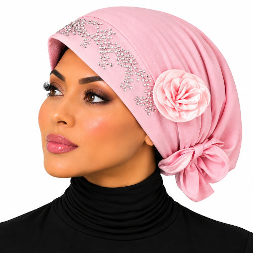 A woman in profile wearing a light pink hijab with silver rhinestone embellishments and a matching pink fabric flower accent, paired with a black turtleneck top against a plain white background.