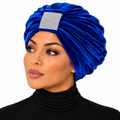 A woman wearing a royal blue velvet turban headwrap with a rhinestone-embellished square accent on the front, paired with a black turtleneck against a plain white background.