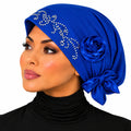 A woman wearing a royal blue satin headscarf with silver rhinestone embellishments, a large fabric rose, and a side bow, paired with a black turtleneck against a white background.