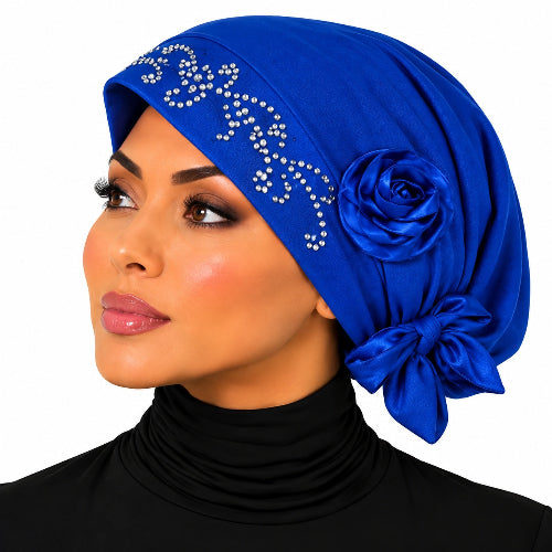 A woman wearing a royal blue satin headscarf with silver rhinestone embellishments, a large fabric rose, and a side bow, paired with a black turtleneck against a white background.